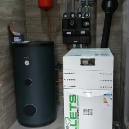 Zestaw urządzeń grzewczych w kotłowni: zasobnik ciepłej wody Kospel, piec na pellet z podajnikiem i zbiornikiem, grupa pompowa i naczynie przeponowe, instalacja w płytkach imitujących drewno.