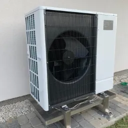 Biała jednostka zewnętrzna na metalowej konstrukcji wsporczej, zainstalowana przy jasnej ścianie budynku, widoczne przewody i narzędzie na kostce brukowej.