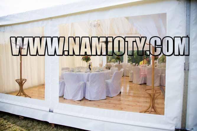 Widok wnętrza białego namiotu bankietowego z drewnianą podłogą, okrągłymi stołami nakrytymi białymi obrusami i krzesłami w pokrowcach, widoczne przez okno namiotu. Na zdjęciu widoczny znak wodny.