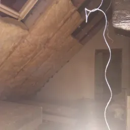 Poddasze w trakcie ocieplania wełną mineralną, widoczne fragmenty drewnianej konstrukcji dachu i okno połaciowe. W tle widoczne nieotynkowane ściany i zwisający przewód.