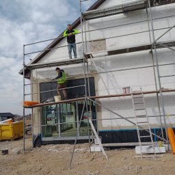 Usługi remontowo-budowlane - Prace na rusztowaniu podczas ocieplania ścian budynku w Biłgoraju. Widoczni dwaj pracownicy w odblaskowych kamizelkach. Ściana z białego styropianu.