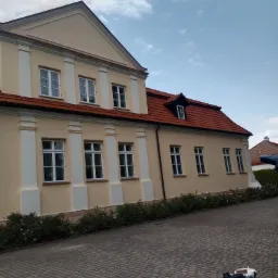 Jasna elewacja budynku z czerwonym dachem, białe kolumny przy oknach. Na brukowanym podjeździe widoczne narzędzia malarskie.