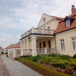Jasna elewacja budynku z balkonem z balustradą, widok z dołu na fasadę z kolumnami i logo na szczycie. Kostka brukowa i zieleń w otoczeniu.