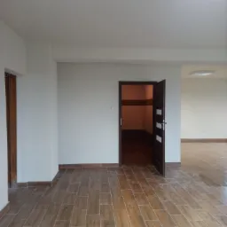 Puste, jasne wnętrze z brązową podłogą imitującą drewno, białe ściany i otwarte drzwi do pomieszczenia z brązowymi płytkami na ścianach.