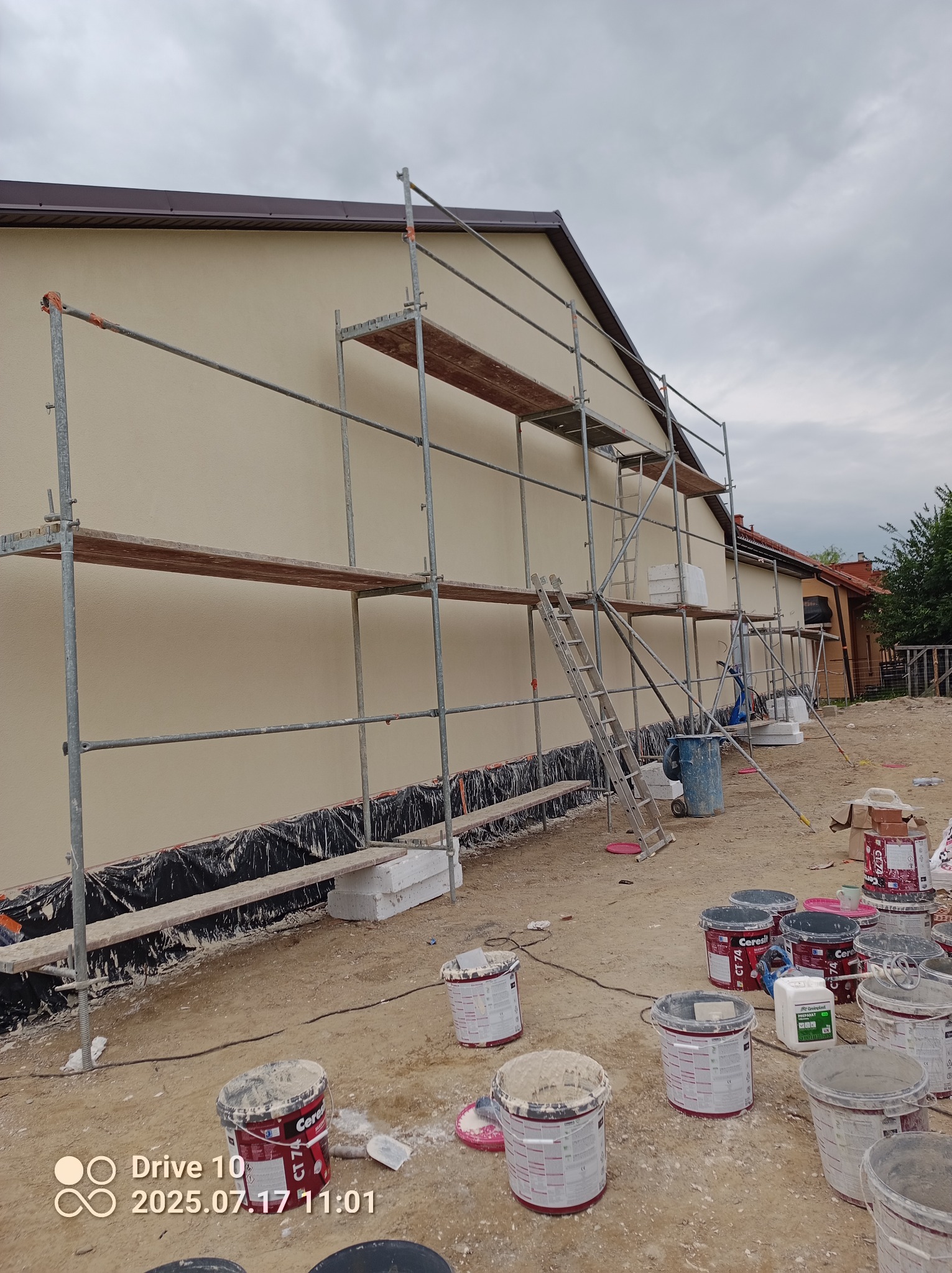 Elewacja budynku w trakcie prac: kremowa ściana, rusztowanie, farby i narzędzia. Widoczne detale wykończenia i przygotowania do malowania.