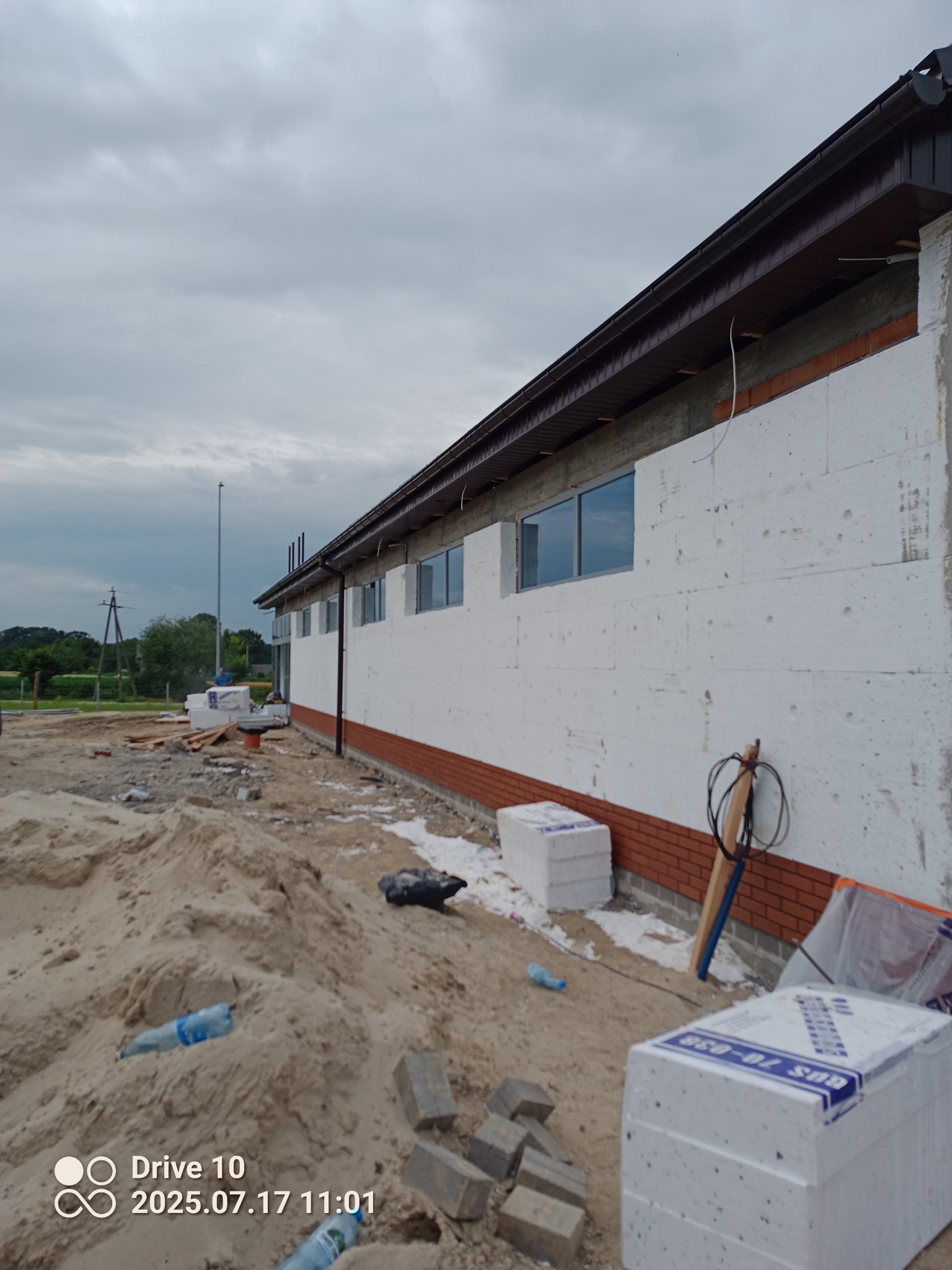 Ocieplanie budynku styropianem: widok na ścianę z białym styropianem, ceglanym cokołem i oknami, z materiałami budowlanymi na pierwszym planie.