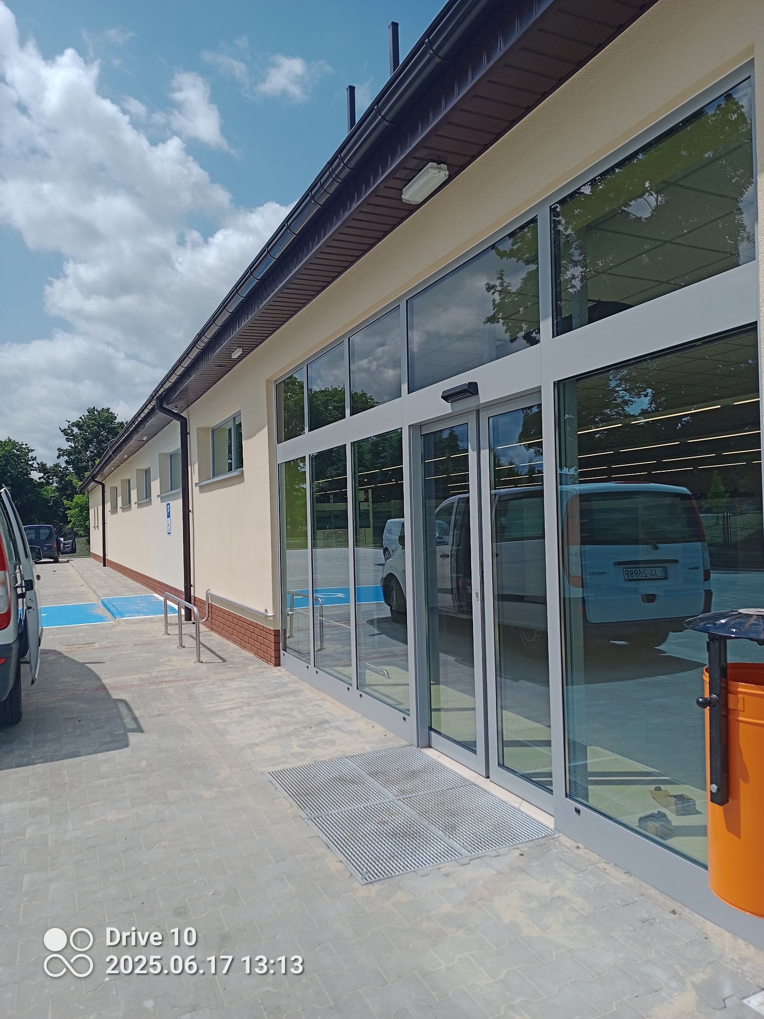 Jasna elewacja budynku z dużymi oknami i automatycznymi drzwiami, widok z zewnątrz. Na chodniku widoczny pomarańczowy kosz na śmieci i zaparkowane samochody.