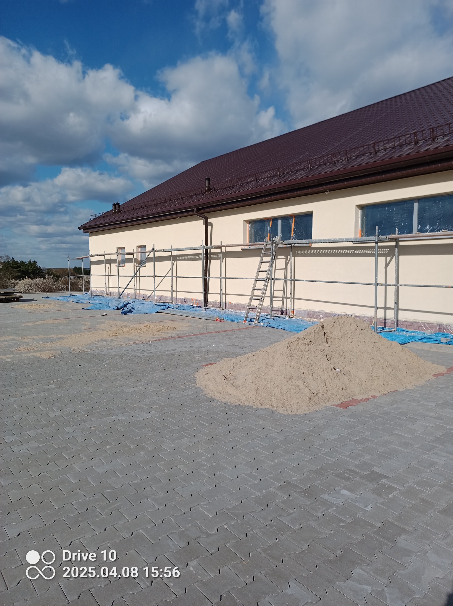 Budynek w trakcie prac elewacyjnych: rusztowania, zabezpieczone okna folią, piasek na kostce brukowej. Widok w pogodny dzień, brązowy dach.
