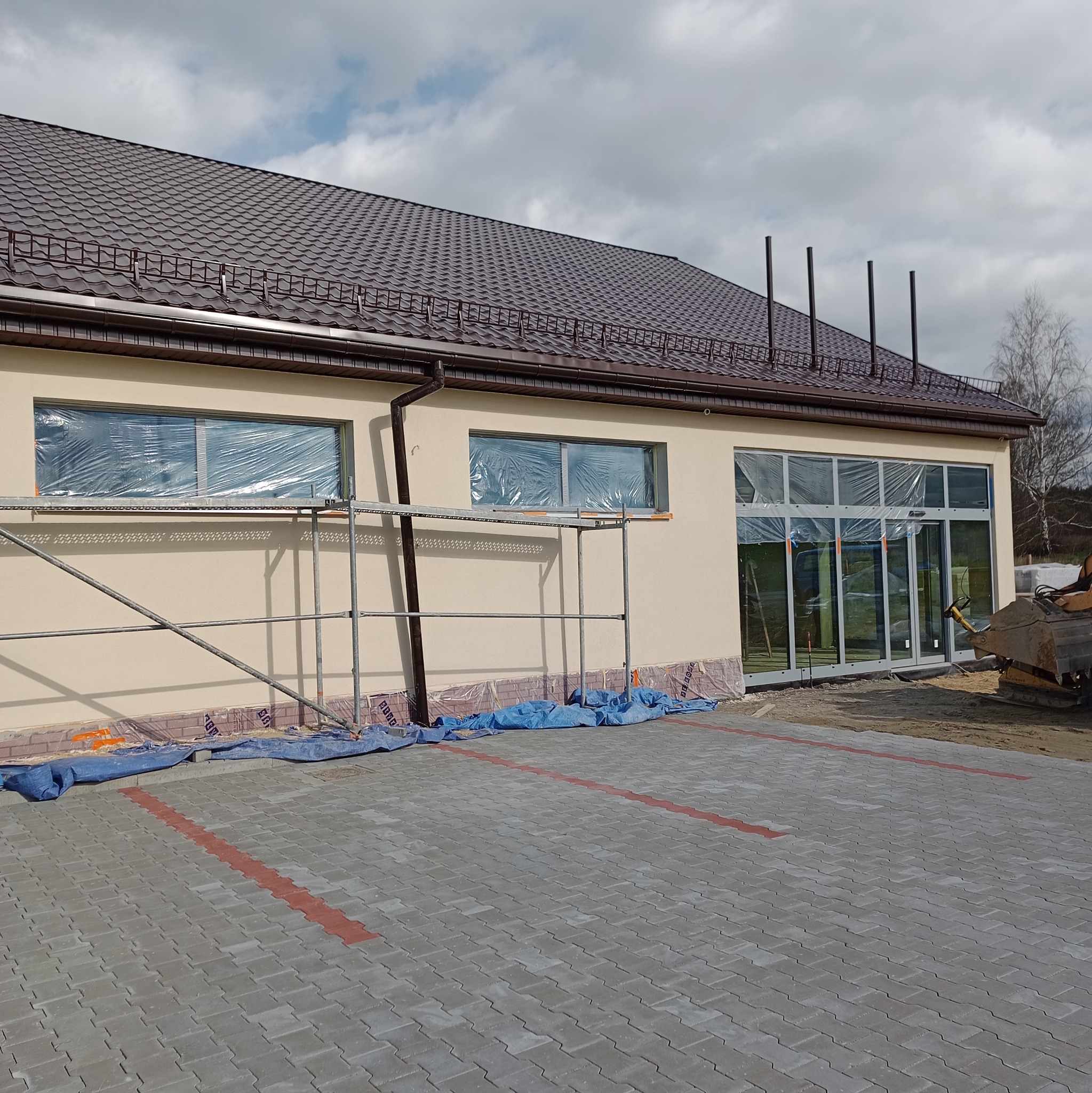 Budynek po tynkowaniu z oknami zabezpieczonymi folią, rusztowaniem i kostką brukową. Widoczne detale wykończenia dachu i rynny. Prace w toku, teren zabezpieczony.