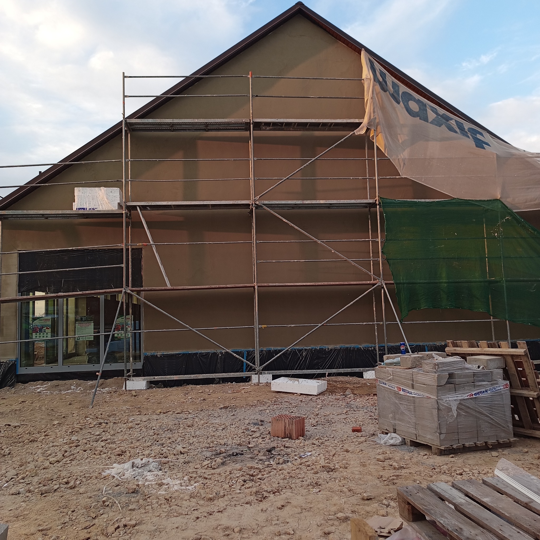 Budynek w trakcie tynkowania elewacji, widoczne rusztowania, materiały budowlane i zabezpieczenia. Częściowo zakryty siatką i folią. Widoczne okna i drzwi.