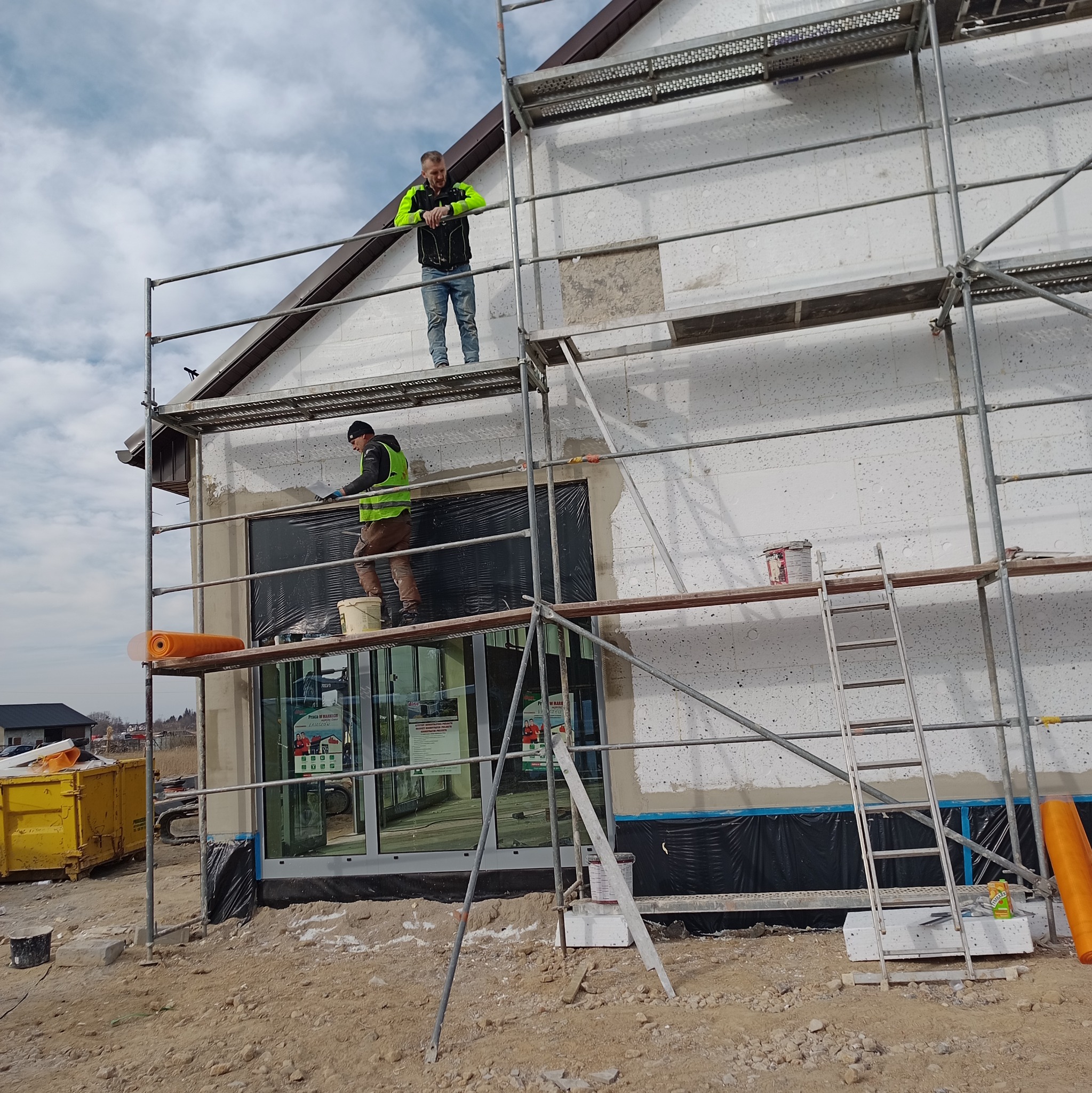 Prace na rusztowaniu podczas ocieplania ścian budynku w Biłgoraju. Widoczni dwaj pracownicy w odblaskowych kamizelkach. Ściana z białego styropianu.