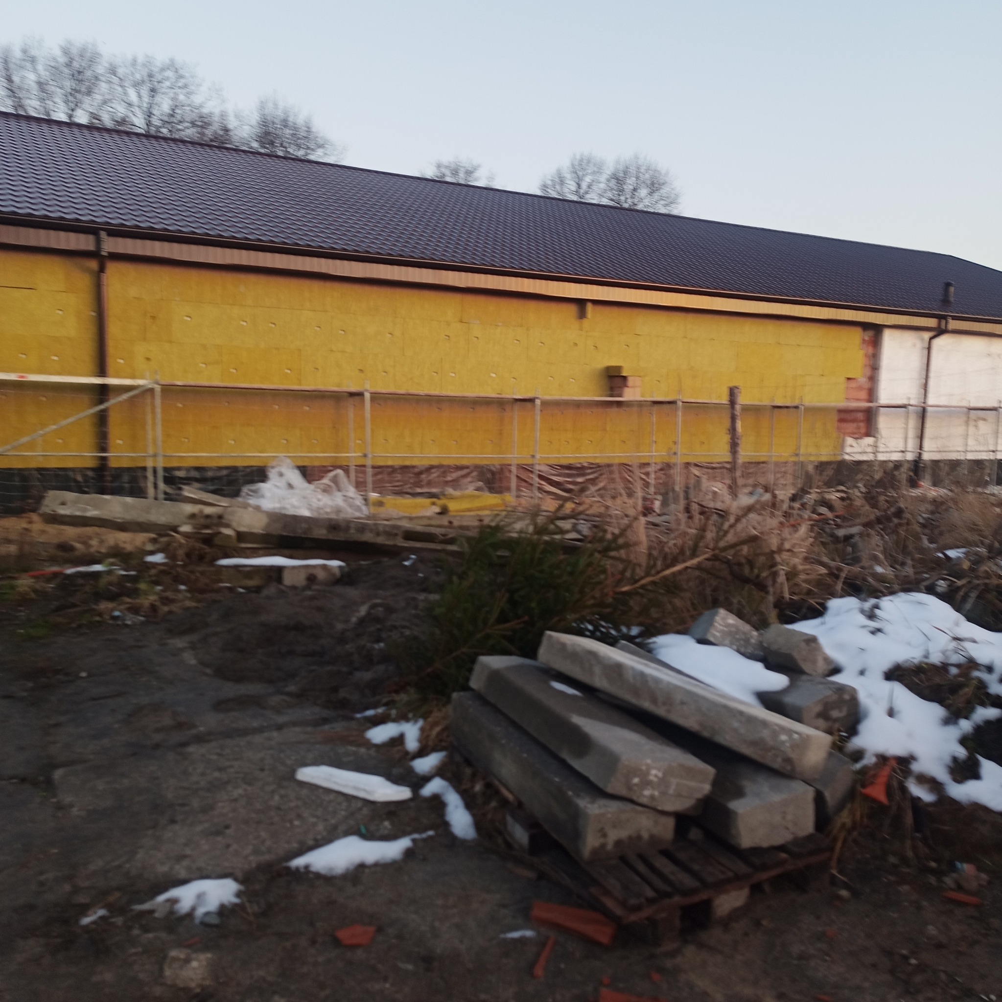 Budynek w trakcie ocieplania: żółta wełna mineralna na elewacji, widoczne rusztowania i materiały budowlane. Zima, śnieg na ziemi.