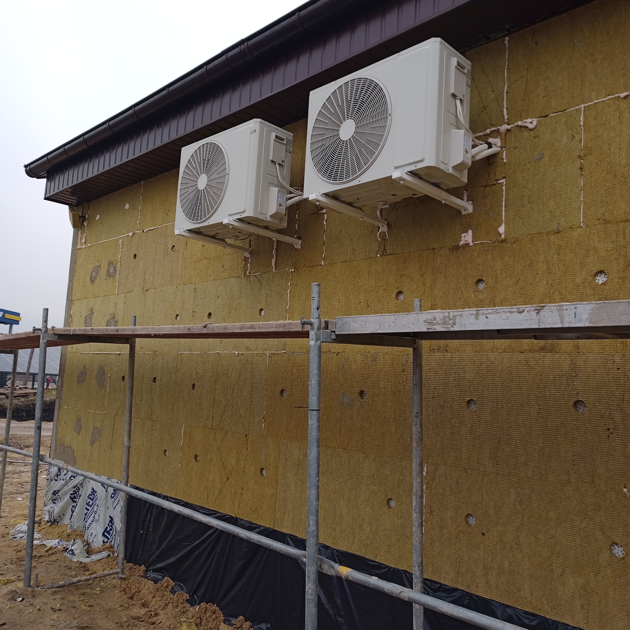 Ocieplona wełną mineralną ściana budynku z zamontowanymi dwoma klimatyzatorami. Widoczne rusztowanie i folia fundamentowa. Etap prac przed nałożeniem tynku.