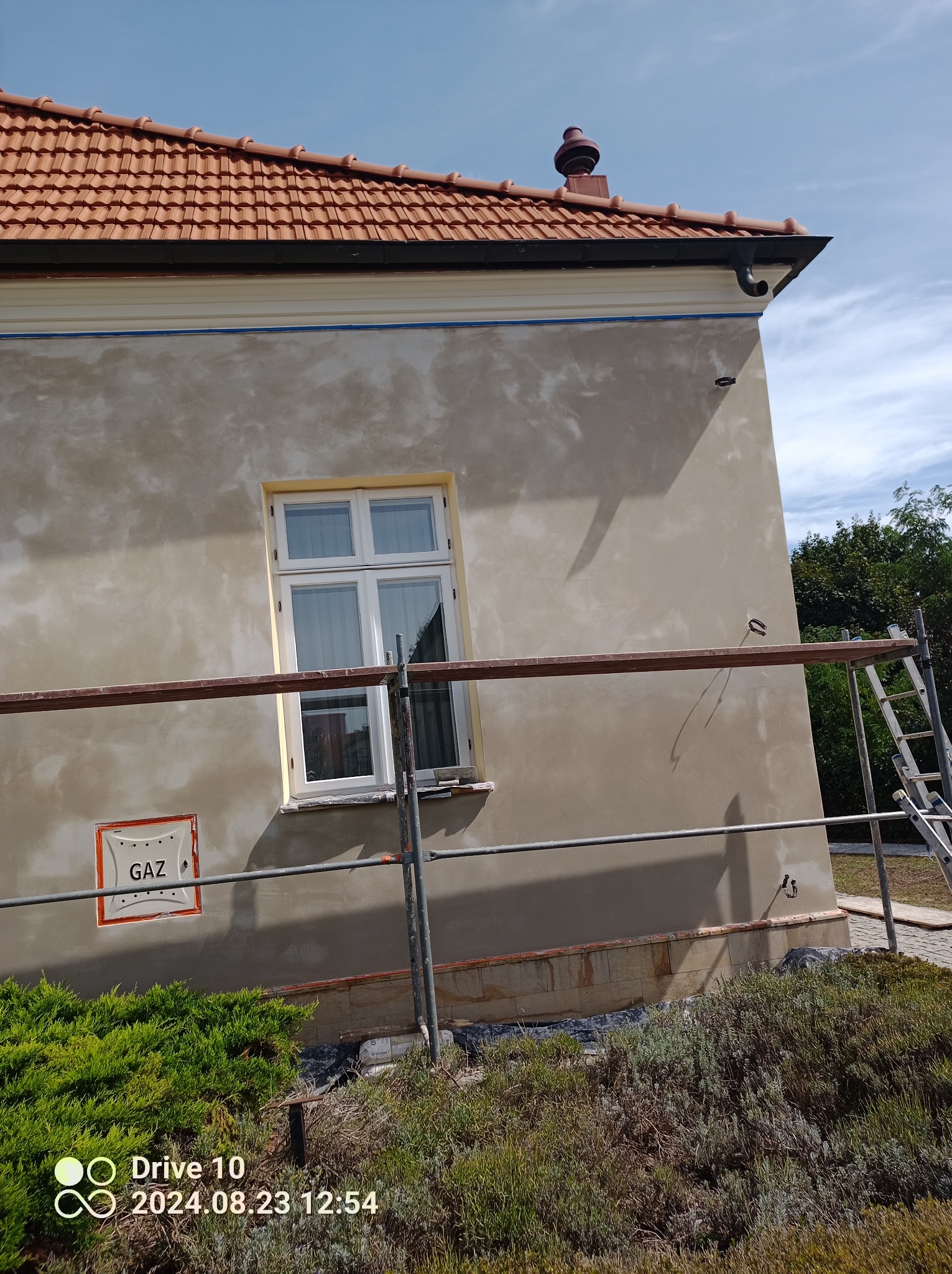 Fragment elewacji budynku w trakcie prac wykończeniowych. Widoczne rusztowanie, okno, znak gazu i surowa struktura tynku. Dach z czerwoną dachówką.