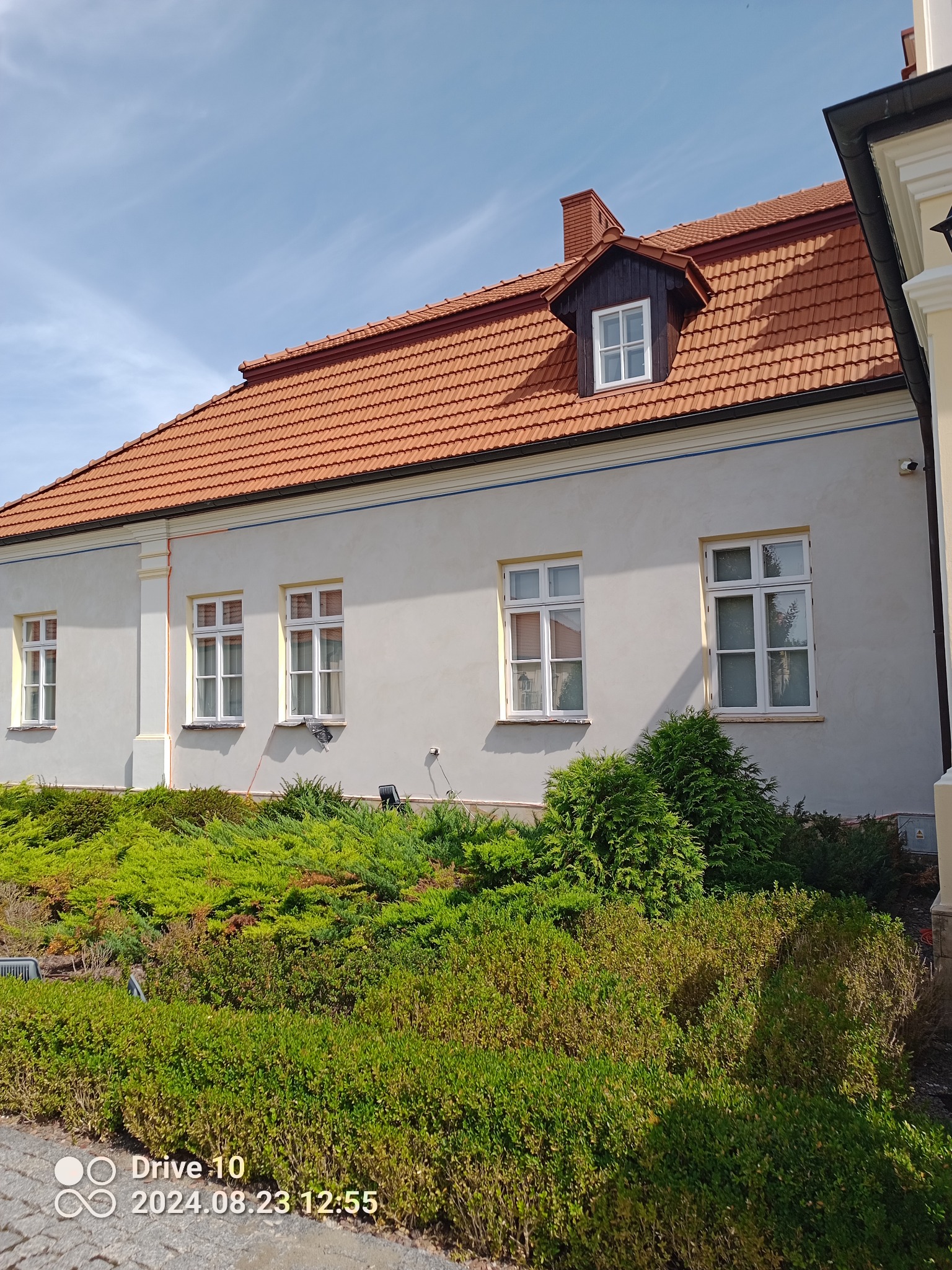 Jasna elewacja budynku z czerwoną dachówką i białymi oknami, otoczona zielenią. Widoczne detale architektoniczne i elementy dekoracyjne.