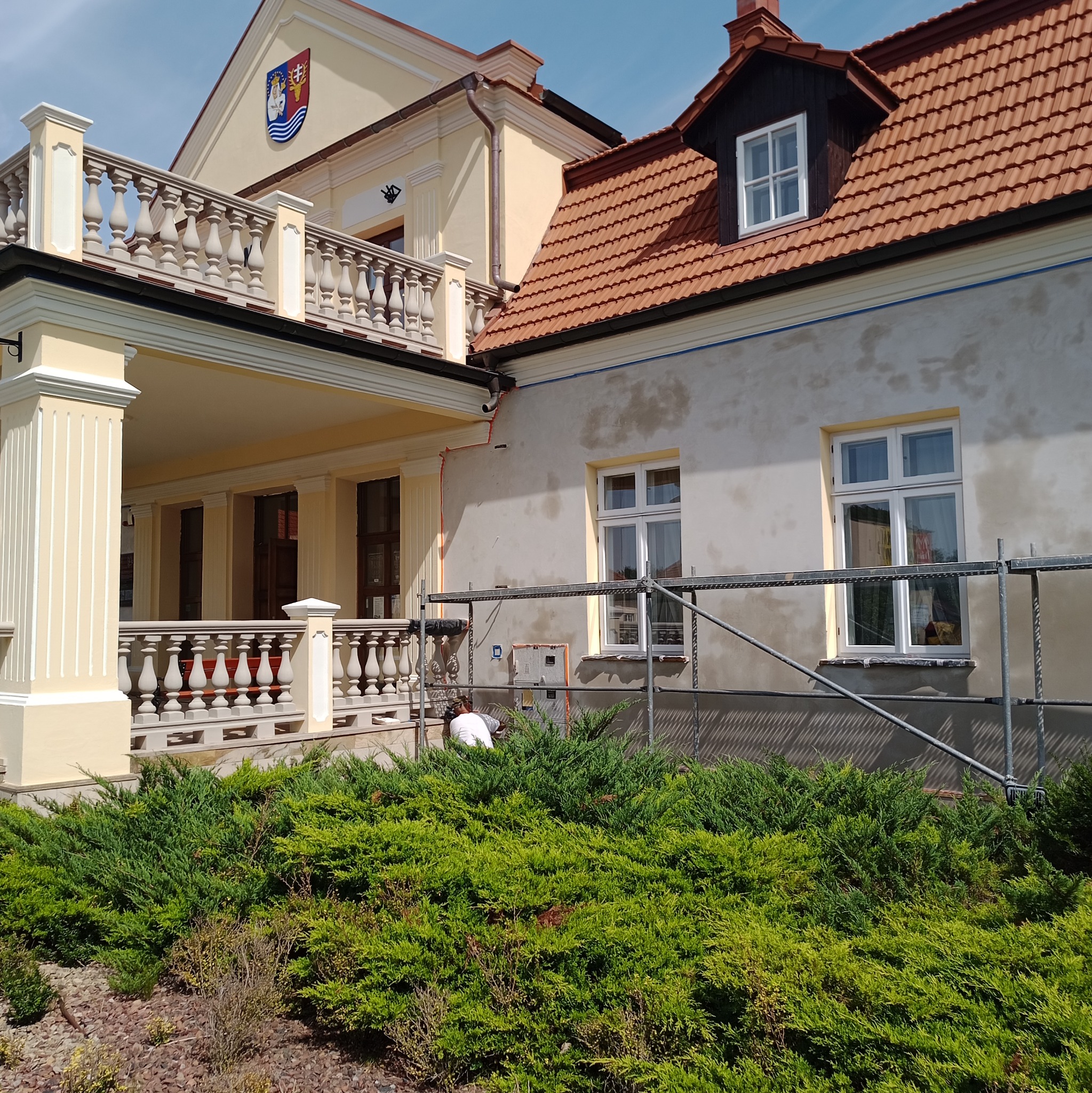 Prace elewacyjne przy budynku z balkonem i herbem. Widoczne rusztowania i pracownik w trakcie szpachlowania/malowania. Częściowo otynkowana elewacja.