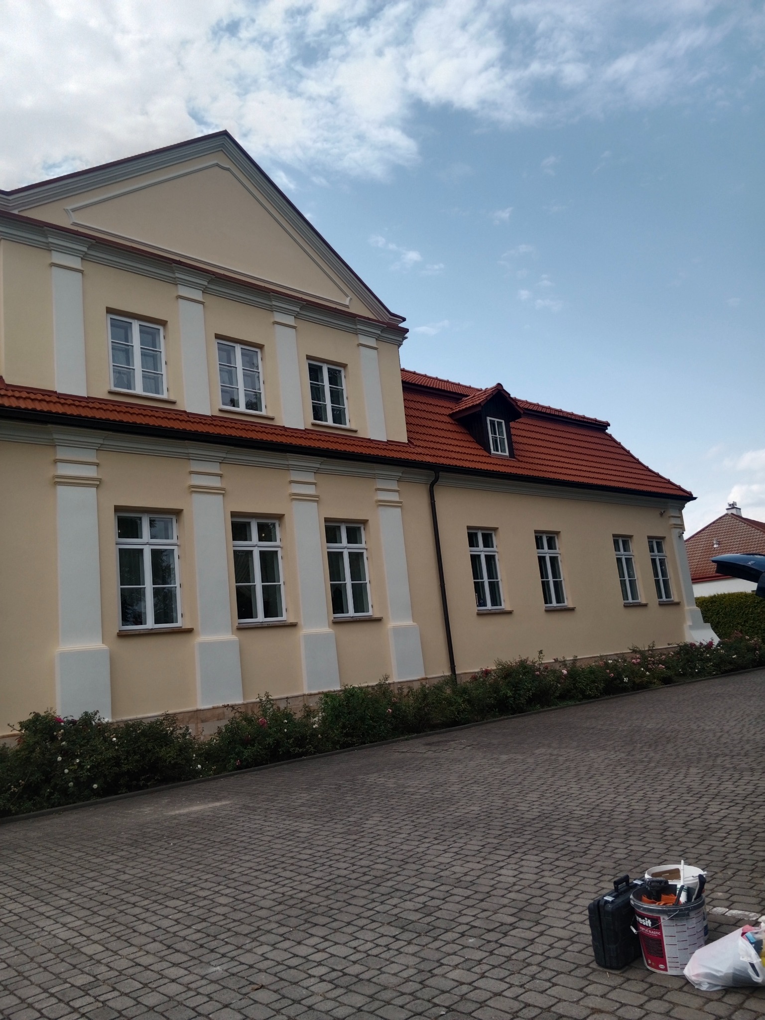 Jasna elewacja budynku z czerwonym dachem, białe kolumny przy oknach. Na brukowanym podjeździe widoczne narzędzia malarskie.