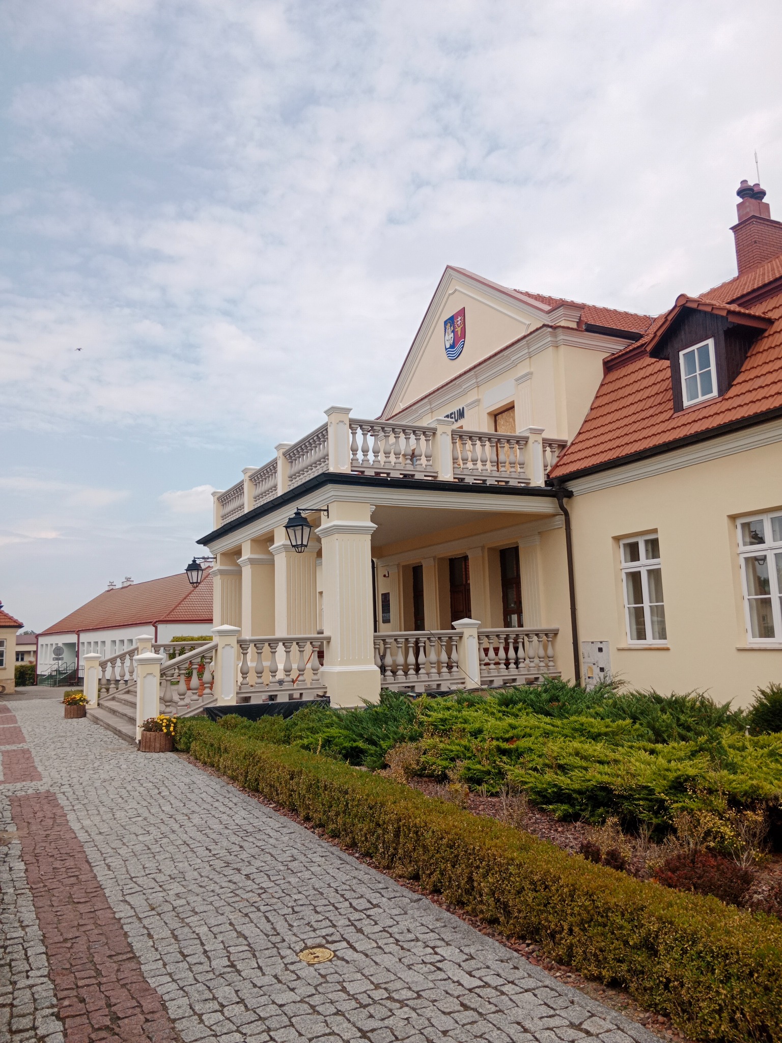 Jasna elewacja budynku z balkonem z balustradą, widok z dołu na fasadę z kolumnami i logo na szczycie. Kostka brukowa i zieleń w otoczeniu.