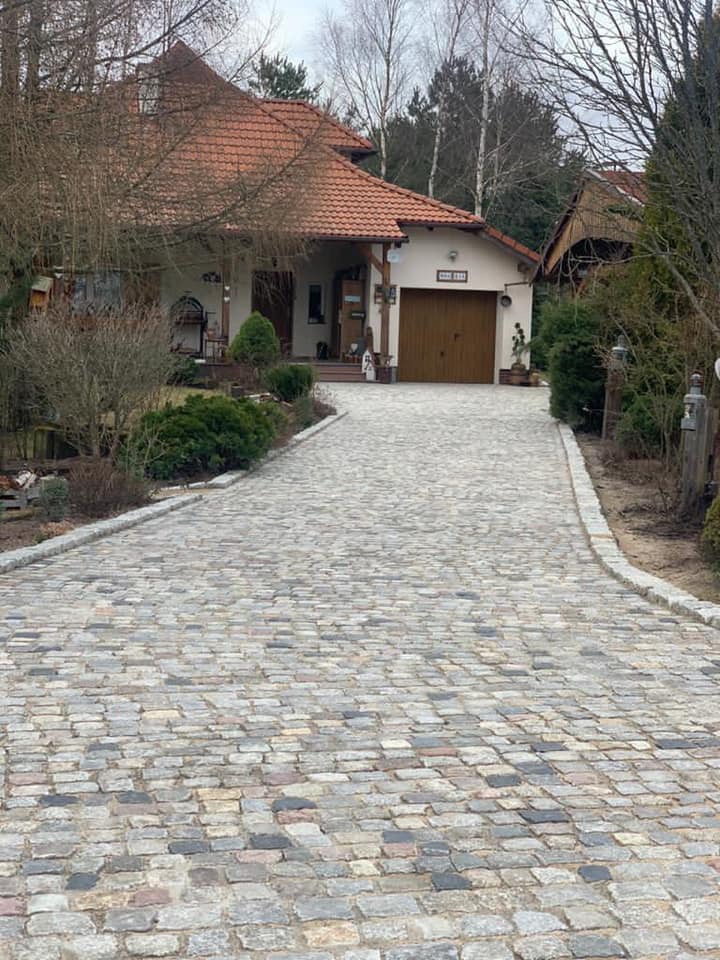 Podjazd do domu wyłożony nieregularną kostką granitową w odcieniach szarości i beżu, obrzeża z jasnej kostki, w tle dom z brązowym dachem i garażem.
