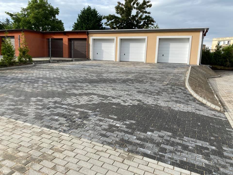 Ułożona kostka brukowa w dwóch odcieniach szarości tworząca wzór na parkingu przed budynkiem z garażami; widoczne zagospodarowanie terenu zielenią i odwodnienie.