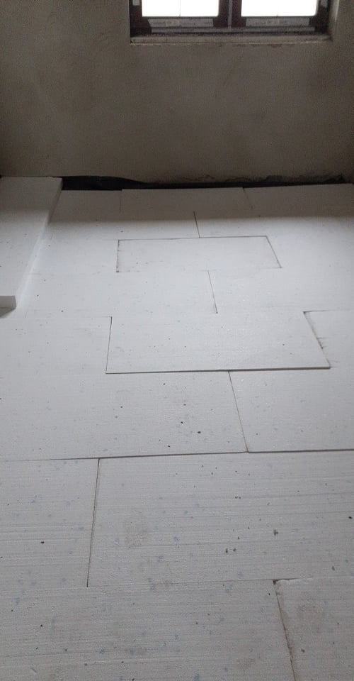 Białe płyty styropianowe ułożone na podłodze w surowym pomieszczeniu z oknem, przygotowane pod instalację ogrzewania podłogowego.