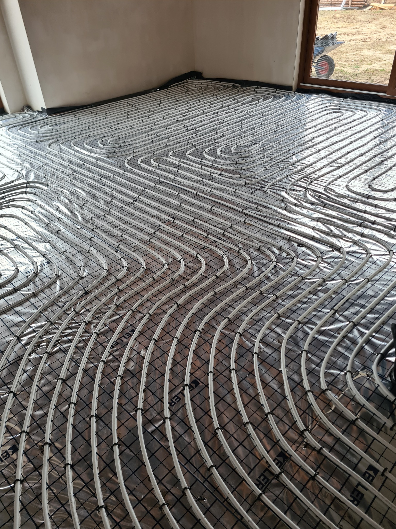 Białe rury ogrzewania podłogowego ułożone na srebrnej folii izolacyjnej w nowym budynku, widoczne czarne klipsy mocujące i fragment okna z widokiem na zewnątrz.