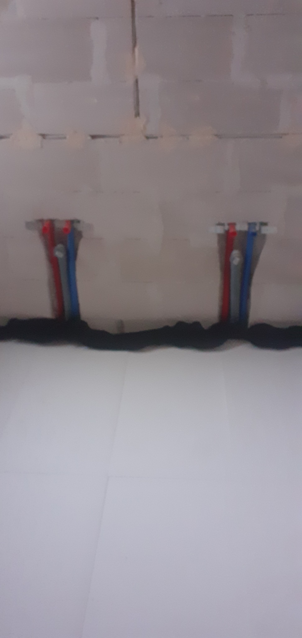 Fragment instalacji podłogowej z rurami PEX w kolorach czerwonym i niebieskim, mocowanymi do ściany z betonu komórkowego, widoczne czarne ocieplenie.