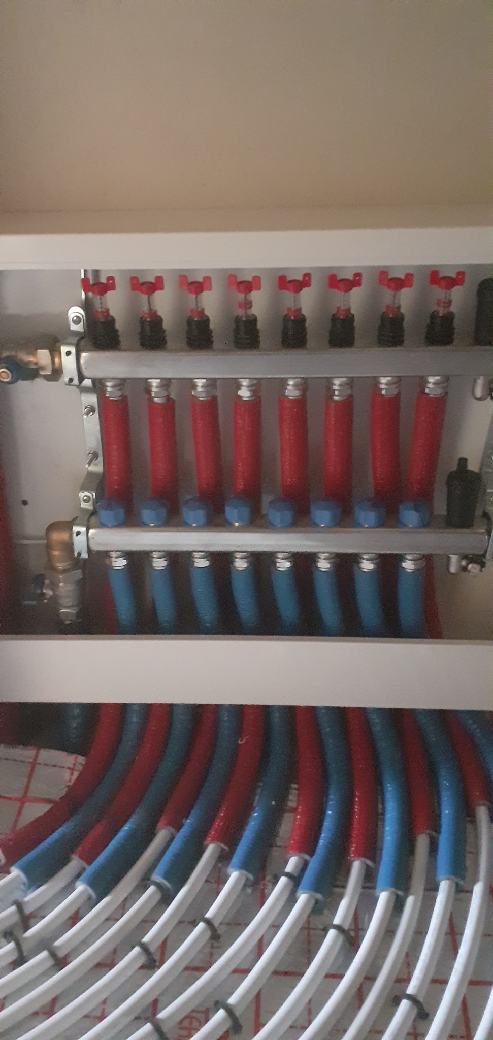 Rozdzielacz ogrzewania podłogowego z czerwonymi i niebieskimi rurami w szafce, widoczne zawory regulacyjne i przyłącza.