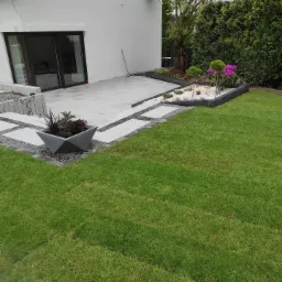 Nowoczesny ogród z geometrycznymi ścieżkami z płyt, rabatą z białym kamieniem i kulistymi drzewkami, minimalistyczna donica z roślinami na tle domu z dużymi oknami przesuwnymi.