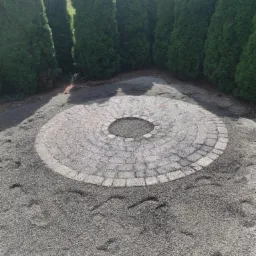 Okrągły plac wyłożony kostką brukową z centralnym otworem, otoczony drobnym żwirem i szpalerem wysokich, zielonych tuj w ogrodzie.