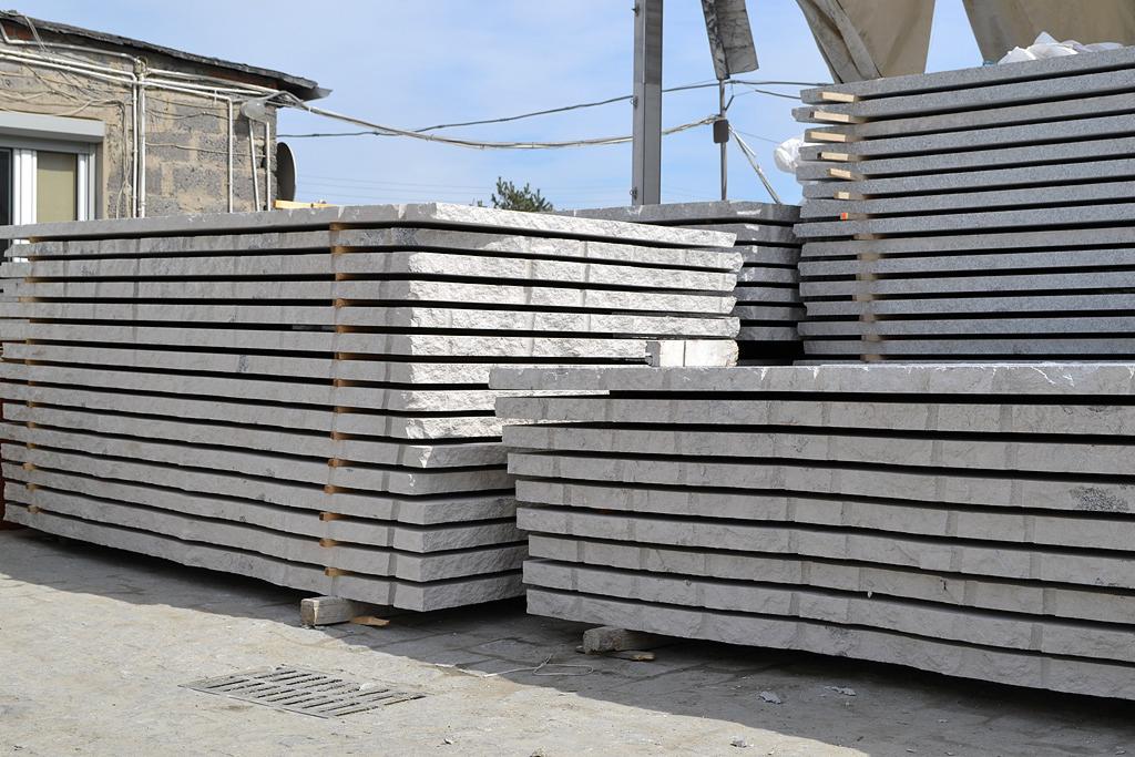 Stosy szarych, obrobionych płyt kamiennych ułożonych na paletach w magazynie, widoczne zarysowania i nierówności na powierzchni kamienia.