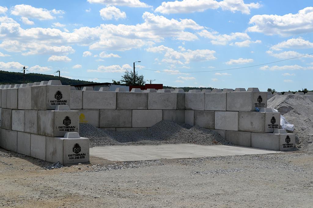 Szare betonowe bloki z logo producenta ułożone w konstrukcję przypominającą mur oporowy, z nasypem żwiru wewnątrz, na tle błękitnego nieba z chmurami.