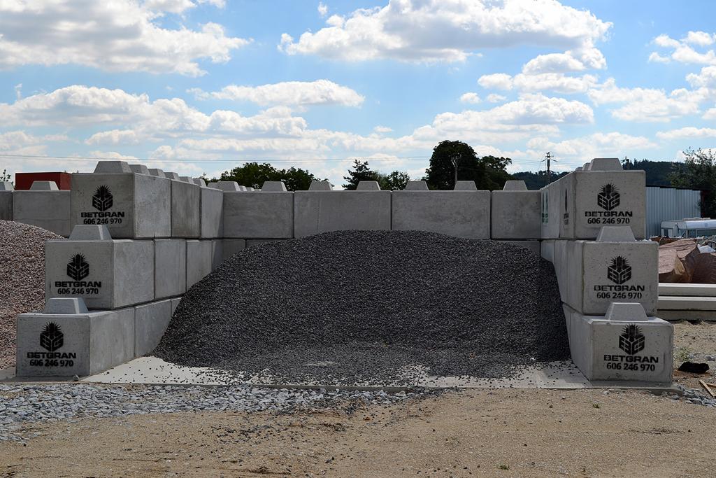 Szare betonowe bloki z nadrukowanym logo 'BETGRAN 606 246 970' ułożone jeden na drugim, tworzące mur oporowy dla nasypu z drobnego, ciemnego kruszywa na placu budowy.