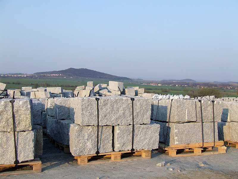 Kostka granitowa w blokach, ułożona na paletach, z widokiem na pagórkowaty krajobraz w tle.