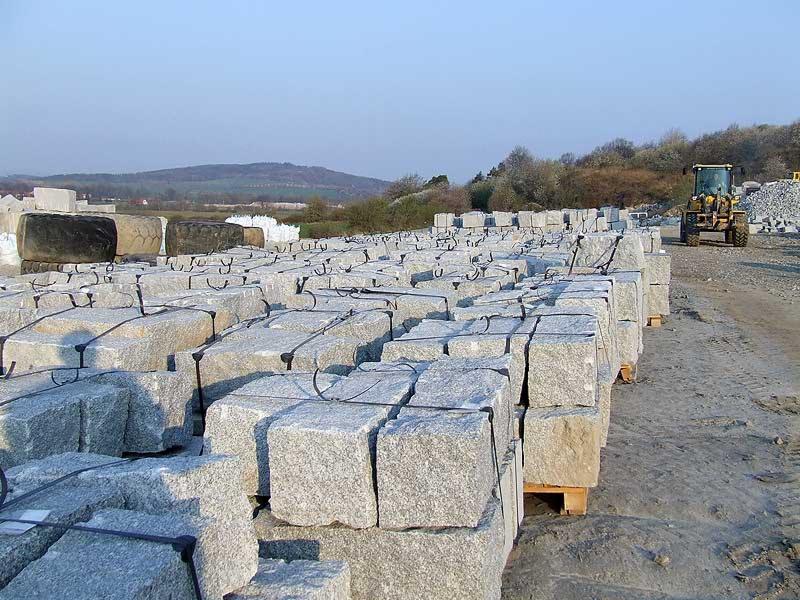 Magazyn kostki granitowej na paletach, w tle koparka i pagórkowaty krajobraz.
