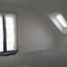 Poddasze w trakcie prac wykończeniowych; okno dachowe i okno pionowe zabezpieczone folią malarską, ściany świeżo pomalowane na biało, widoczne ślady farby na podłodze.
