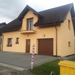 USŁUGI OGÓLNOBUDOWLANE TERLECKI - Żółty dom z ciemnobrązowym dachem i garażem, szara kostka brukowa na podjeździe, widoczne kwiaty w oknach, pochmurne niebo.