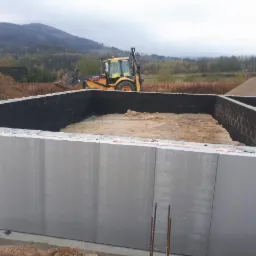 Wykop pod fundamenty zabezpieczony izolacją, z widocznym traktorem Volvo w tle i stertą ziemi.
