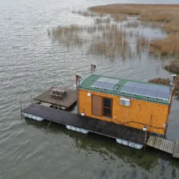 Drewniany pływający domek z panelami słonecznymi na dachu i klimatyzacją na ścianie, zacumowany do pomostu na jeziorze w pochmurny dzień.
