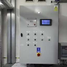Szara szafa sterownicza z wyświetlaczem, przyciskami i lampkami kontrolnymi, zamontowana na ścianie w pomieszczeniu technicznym, obok izolowanych rur i metalowych szyn montażowych.