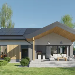 Nowoczesny parterowy dom z panelami słonecznymi na dachu, częściowo wykończony drewnem, z zadaszonym tarasem i meblami ogrodowymi, otoczony zielenią i niskimi krzewami.