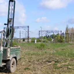 Zabrudzony, ciemnozielony samochód terenowy z wiertnicą na łące w pobliżu linii kolejowej. Wiertło leży na ziemi. Błękitne niebo z chmurami w tle.