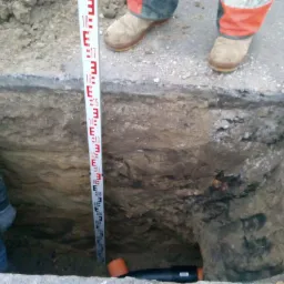 Głęboki wykop w ziemi z widoczną łatą niwelacyjną, pomarańczową rurą i fragmentem instalacji w dole, obok betonowego krawężnika i stóp robotnika w roboczych butach.