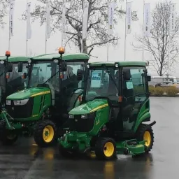 Rząd zielonych traktorów John Deere z żółtymi felgami na placu wystawowym w pochmurny dzień. W tle widoczne flagi i sylwetka mężczyzny.