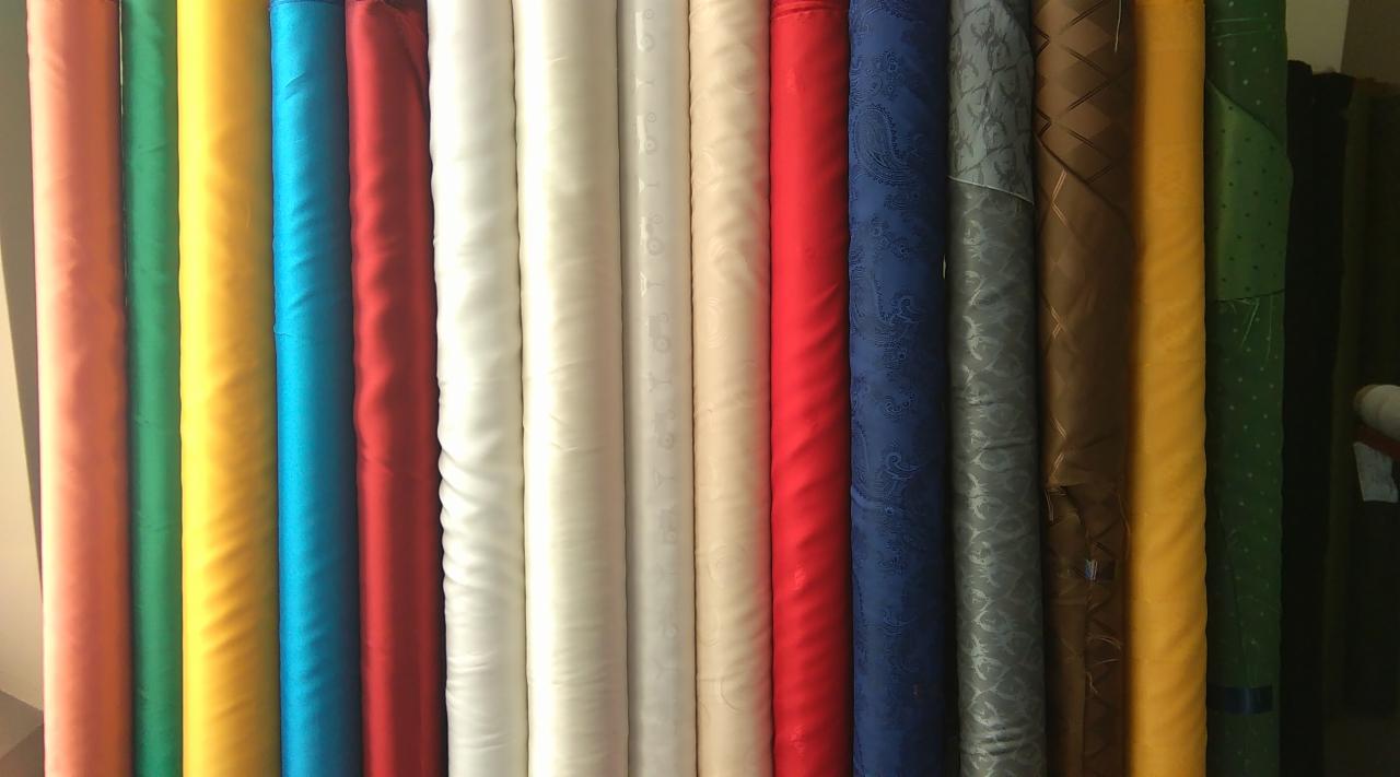 Rzędy kolorowych bel materiałów, w tym tkaniny z wzorami paisley, kratą i kropkami, ustawione pionowo w magazynie.
