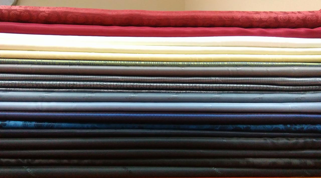 Stos złożonych bel materiałów o różnorodnych wzorach i kolorach, od czerwieni z delikatnym deseniem po ciemne odcienie granatu i brązu, ułożonych równo na sobie.