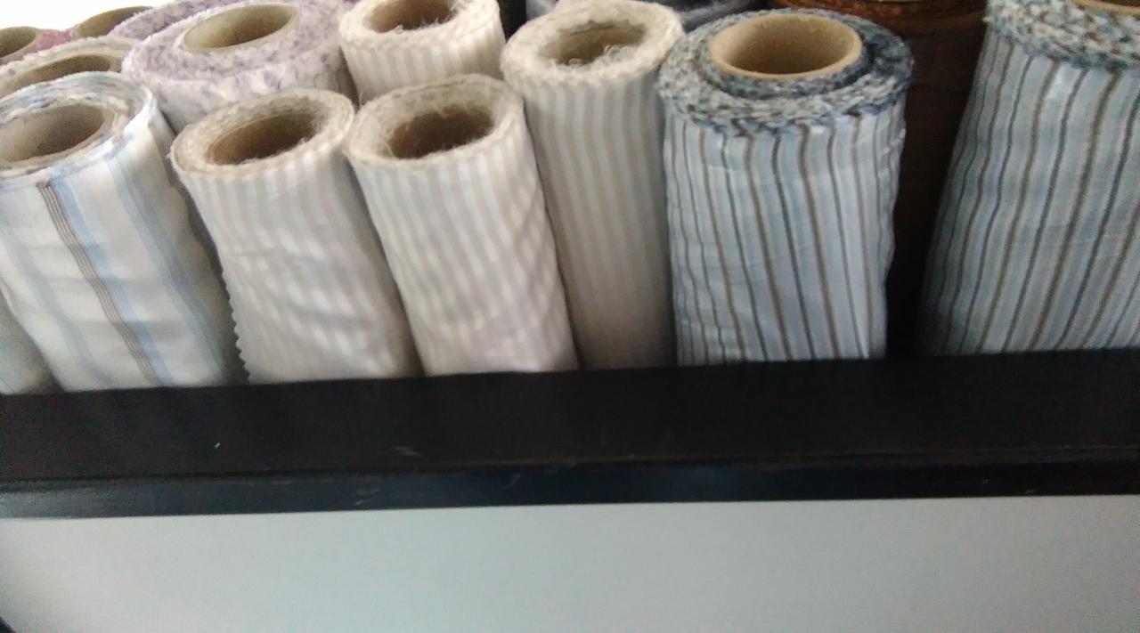 Rzędy rolek tkanin w paski o różnych odcieniach, umieszczone na ciemnej półce w magazynie.