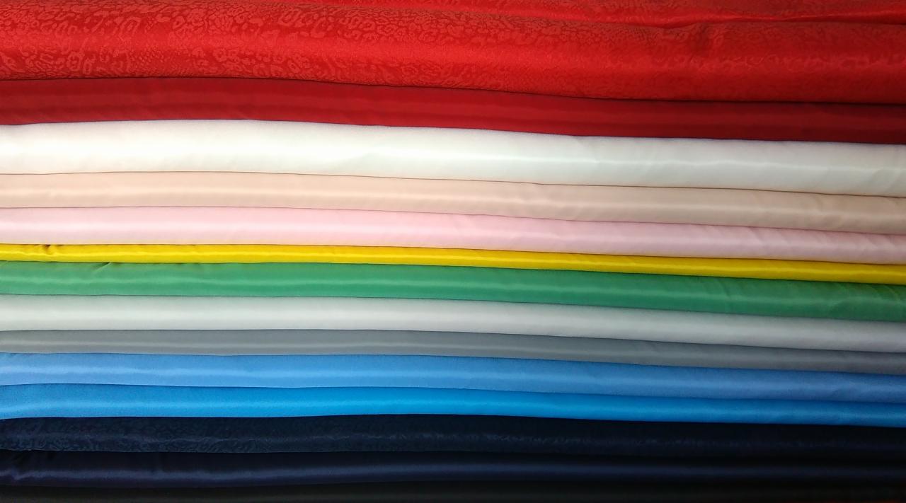 Stos kolorowych tkanin, od czerwonej z delikatnym wzorem po czarną, ułożonych jedna na drugiej, ukazujących różnorodność materiałów.