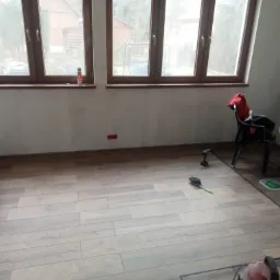 Ułożone panele podłogowe w trakcie prac wykończeniowych, widoczne narzędzia, odkurzacz i szczotka do zamiatania, fragment ściany z niedokończonymi płytkami w jodełkę oraz okna z widokiem na zewnątrz.