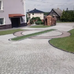 Ułożona kostka granitowa na podjeździe z elementami trawy w kształcie okręgów i linii, brama wjazdowa z figurami orłów, budynek mieszkalny w tle.